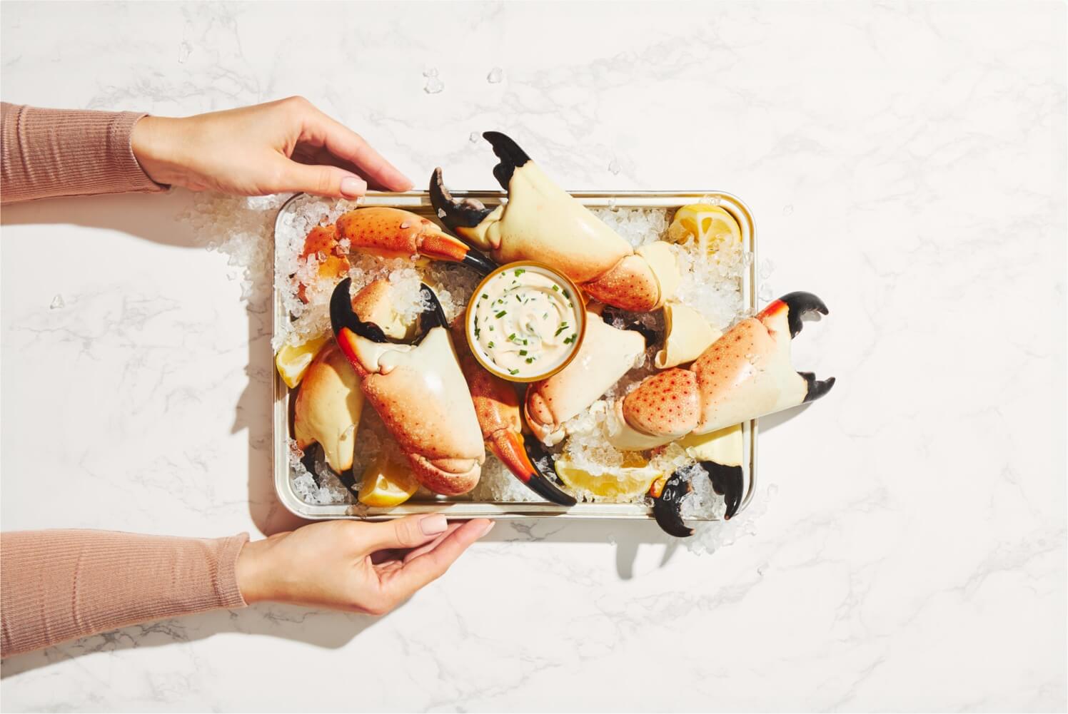 Person holding a tray of cooked crab legs with a small bowl of sauce on a white background