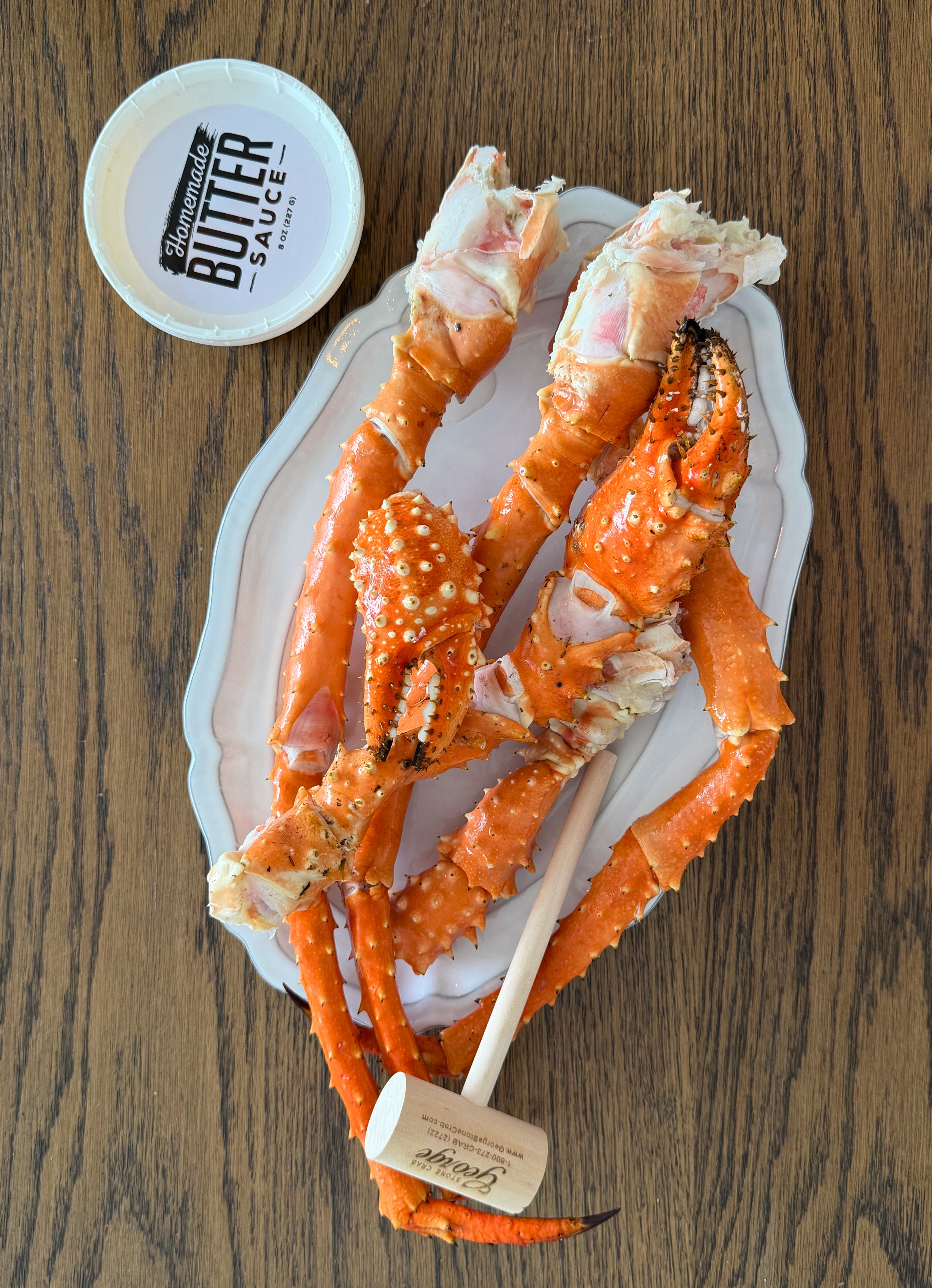 Alaska King Crab legs on a white plate with a wooden mallet and 'Butter Sauce' container on a wooden surface.
